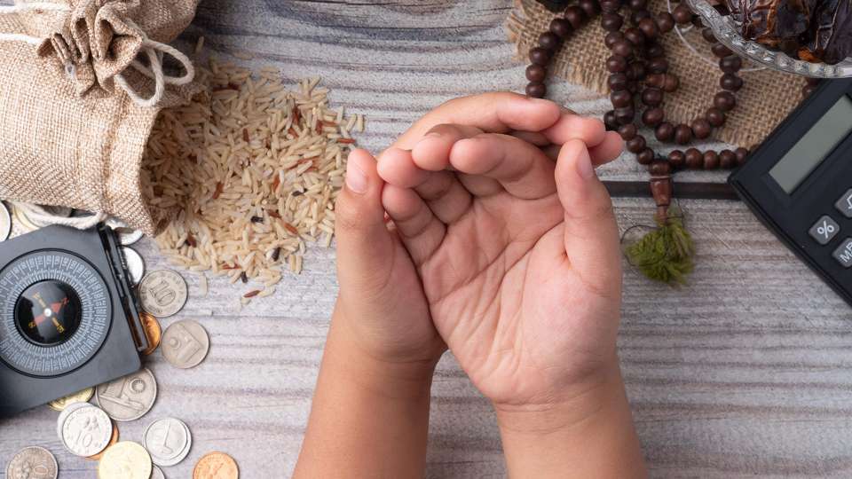Image of child's hands praying over table with rice, prayer compass, coins, prayer beads, dates and calculator.