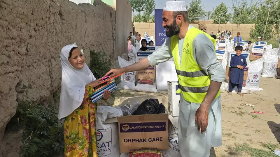 A smiling Afghan orphan girl receives notebooks thanks to your support through Zakat Foundation of America, empowering her education and future.